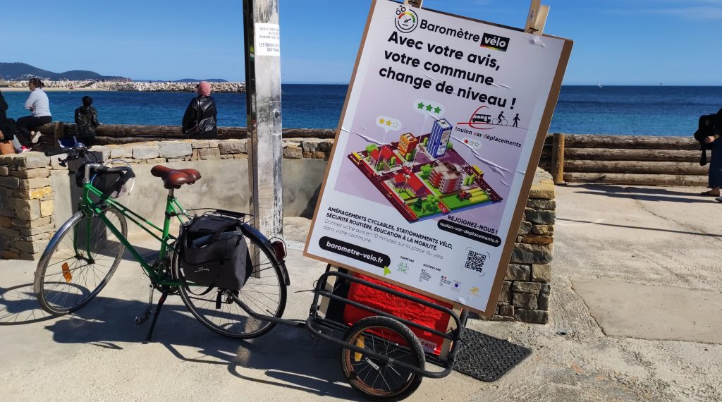 Photo d'un petit vélo vert de ville, avec saccorche et remorque, garé à la Plage du Mourrillon. La remorque porte une énorme affiche (format A0) avec le visuel du baromètre vélo, ainsi que lien et logo de TVD.